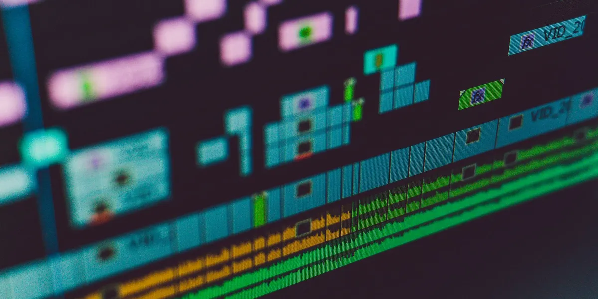 Close-up of an editing timeline on a monitor: stacked violet and teal video clips above amber and green audio waveform tracks, in shallow focus.